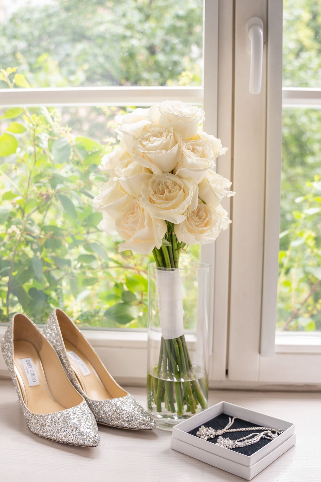 Bukiet białych róż obok brokatowych szpilek ślubnych i biżuterii