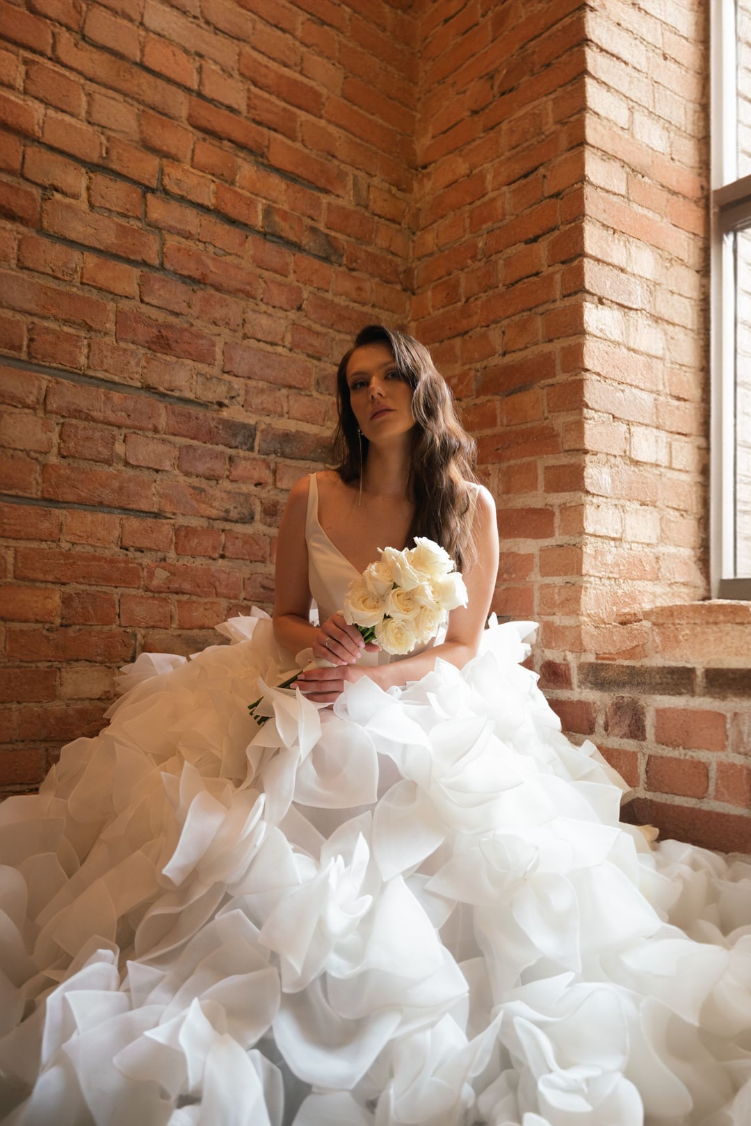 Panna młoda w obszernej sukni siedząca przy ceglastej ścianie z bukietem białych róż