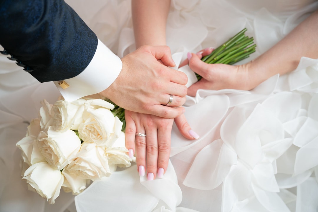 Dłonie pary młodej z obrączkami, bukiet białych róż na tle białej sukni