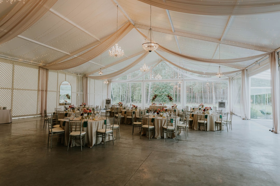 Wnętrze namiotu weselnego z dekoracjami sali weselnej, okrągłymi stołami zastawionymi obrusami w jasnych tonach, krzesłami, kwiatowymi kompozycjami, żyrandolami i półprzezroczystym sufitem z draperiami