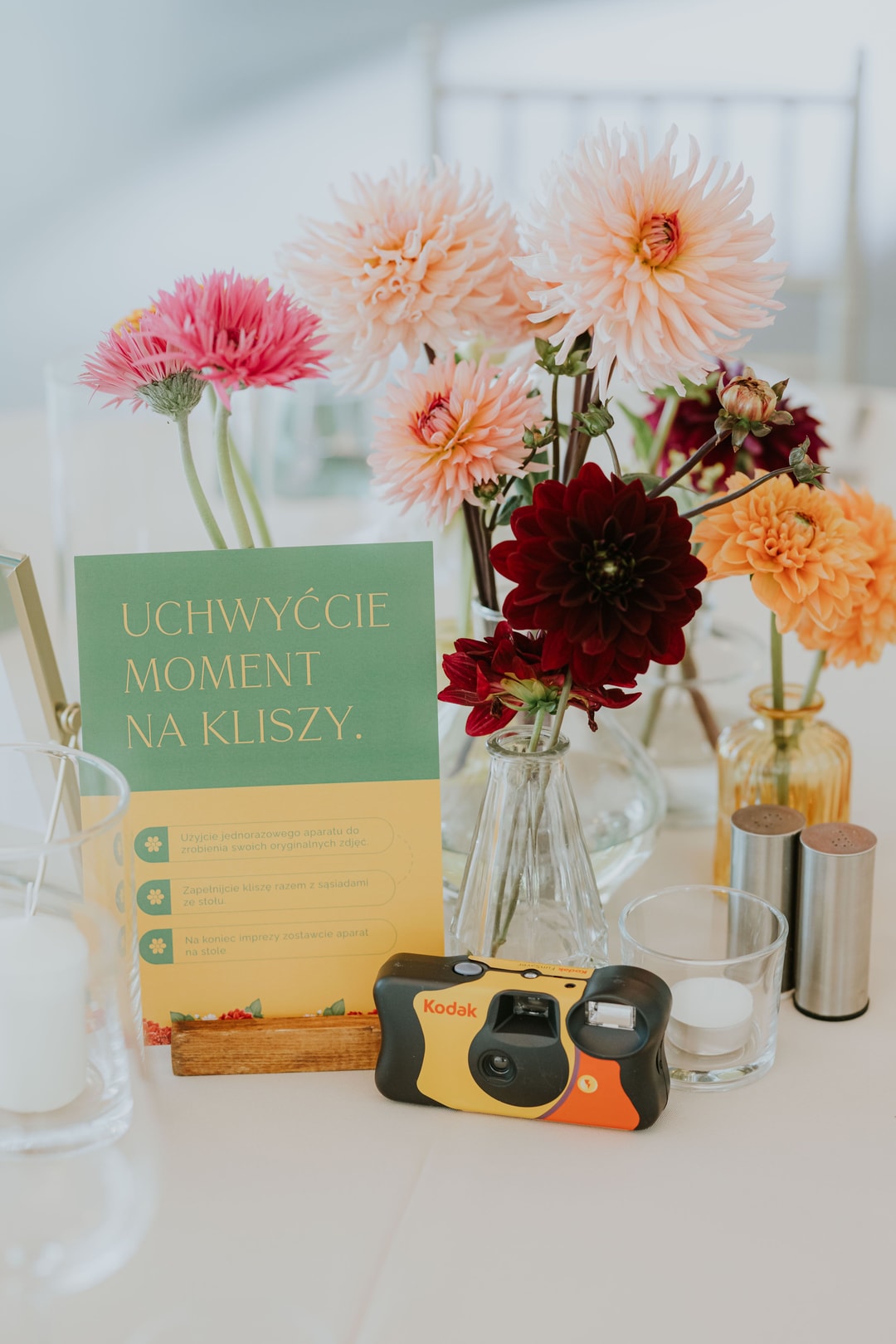 Kompozycja kwiatowa na stół z różowymi i pomarańczowymi dalią, ciemnoczerwonymi kwiatami, zielenią oraz papeterią i aparatem fotograficznym.