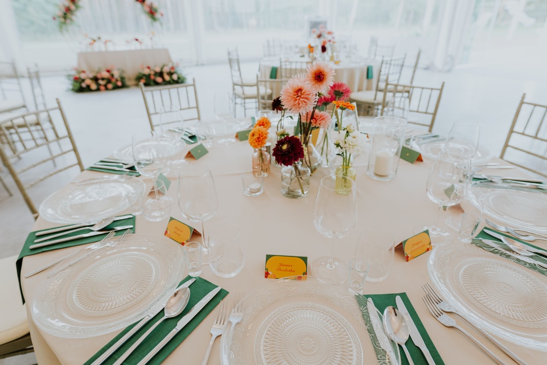 Aranżacja stołu weselnego z białym obrusem, zielonymi serwetkami, talerzami, sztućcami i kompozycją kwiatową w jasnych i pomarańczowych tonach