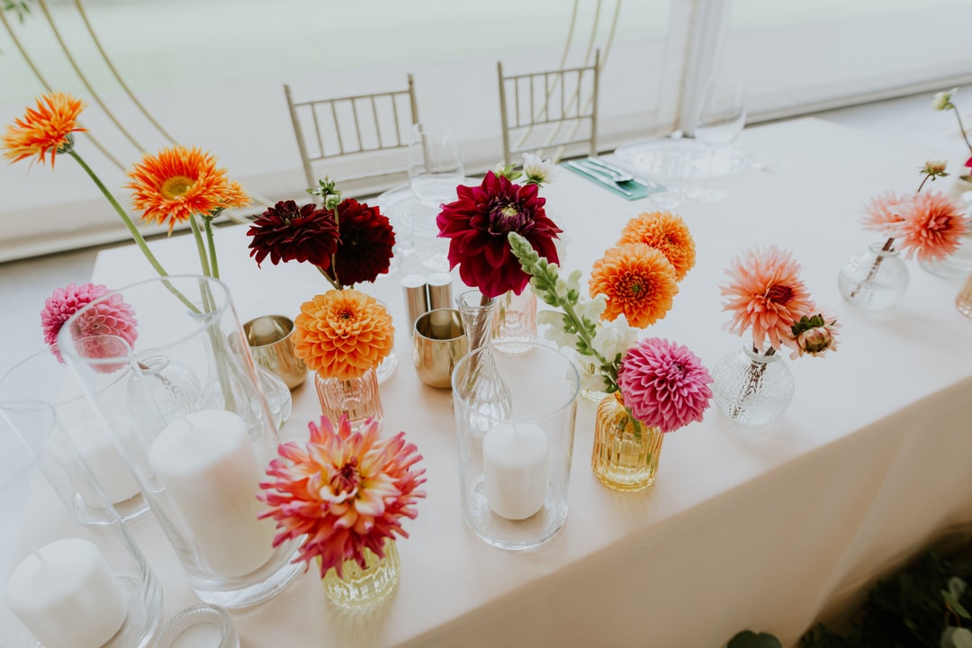 Dekoracja stołu z kompozycjami kwiatowymi w szklanych wazonach. Kwiaty w kolorach pomarańczowym, różowym, bordowym i jasnych tonach z białymi świeczkami.