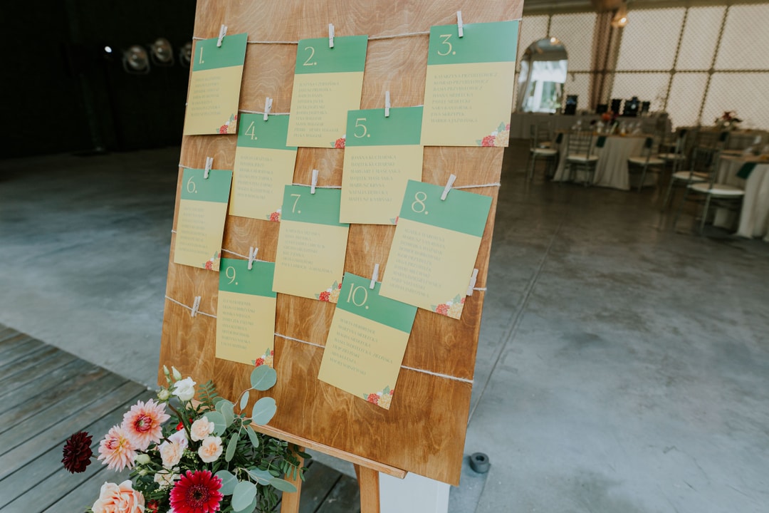 Drewniana tablica z numerami stołów 1-10 na zielonych i kremowych kartach, przypiętych spinaczami, z bukietem kwiatów u podstawy