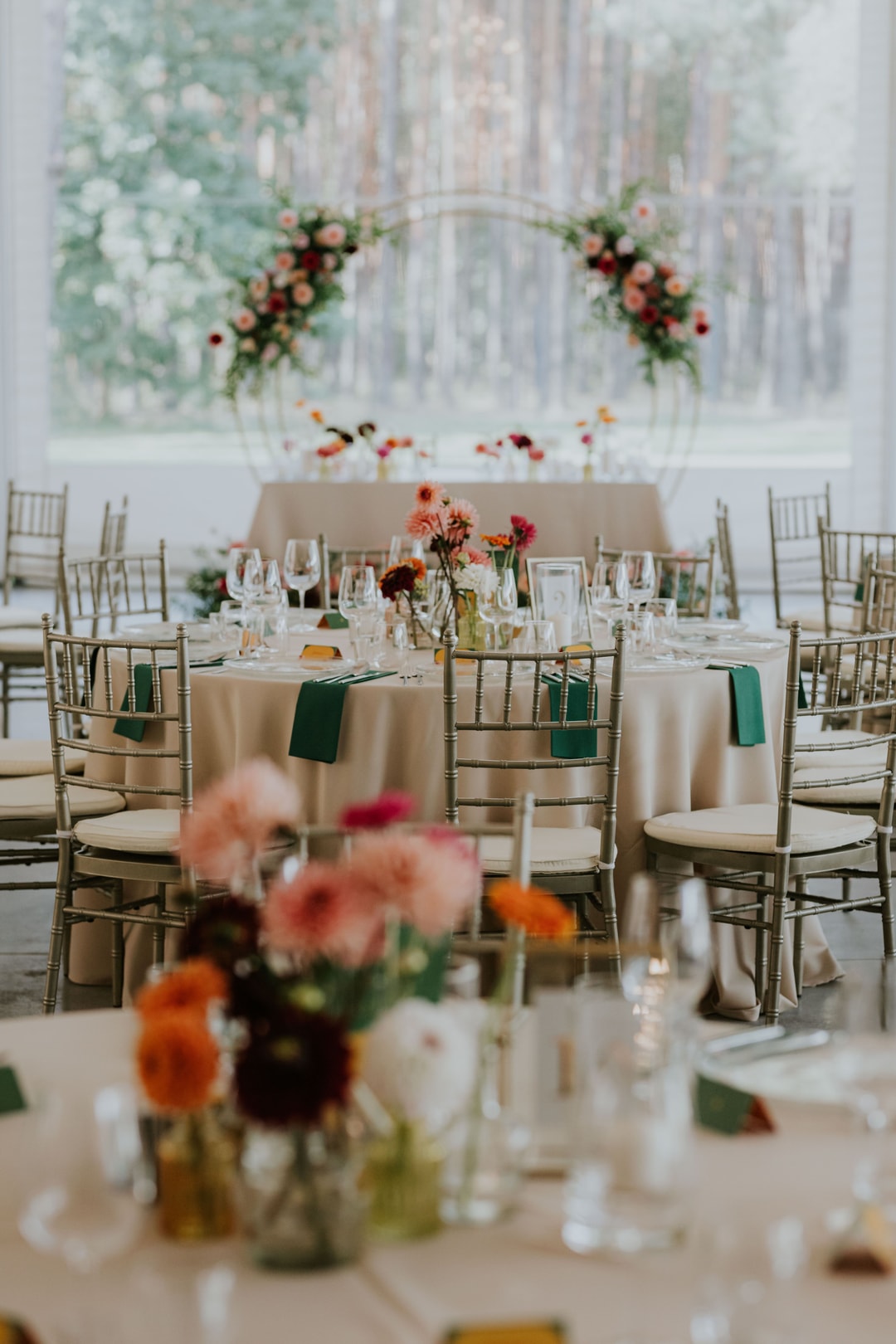 Aranżacja stołów weselnych z beżowymi obrusami, złotymi krzesłami, kompozycjami kwiatowymi w ciepłych tonach i zielonymi serwetkami