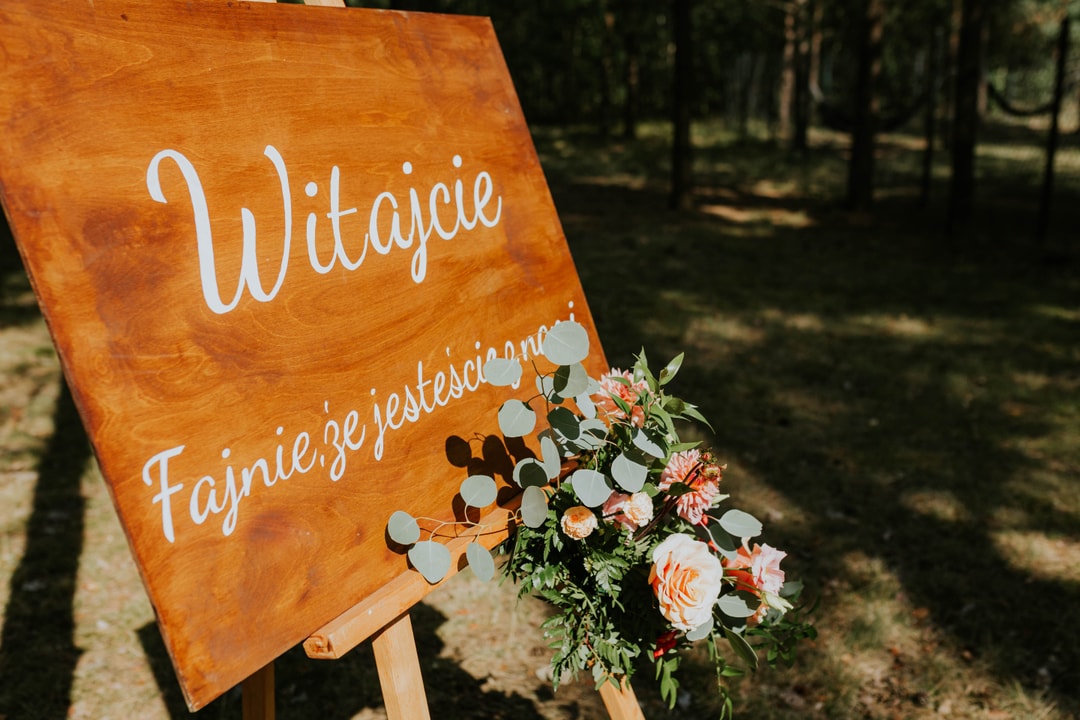 Pomarańczowa tablica z napisem "Witajcie" i "Fajnie się jestęścianie" oraz букиет kwiatowy w ciepłych tonach przy wejściu w lesie