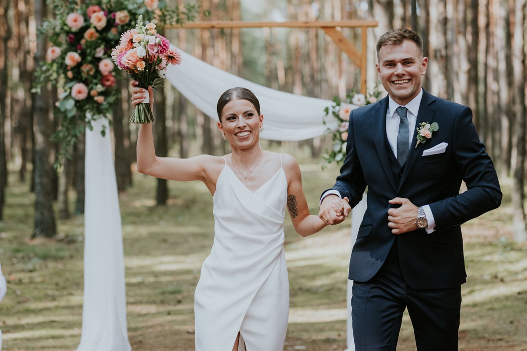 Para Młoda w alejce między drewnianymi słupkami, kobieta trzyma bukiet ślubny w jasnej palecie, mężczyzna w ciemnym garniturze, dekoracja łuku kwiatowego w tle