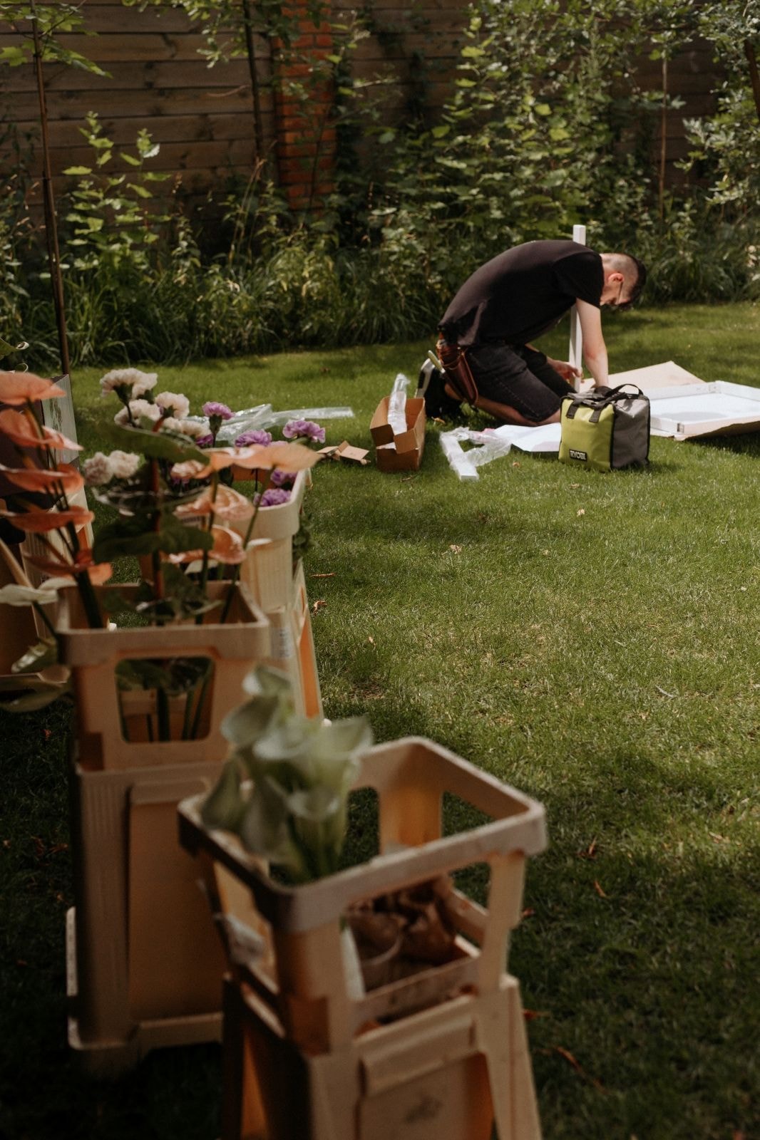 Dekorator ślubny w plenerze: kompozycje kwiatowe w drewnianych skrzynkach na zielonej trawie.