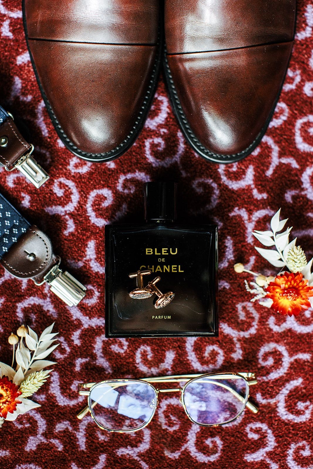 Skórzane brązowe buty, perfumy Bleu de Chanel, okulary przeciwsłoneczne i akcesoria na czerwonym dywanie z ozdobnym wzorem.