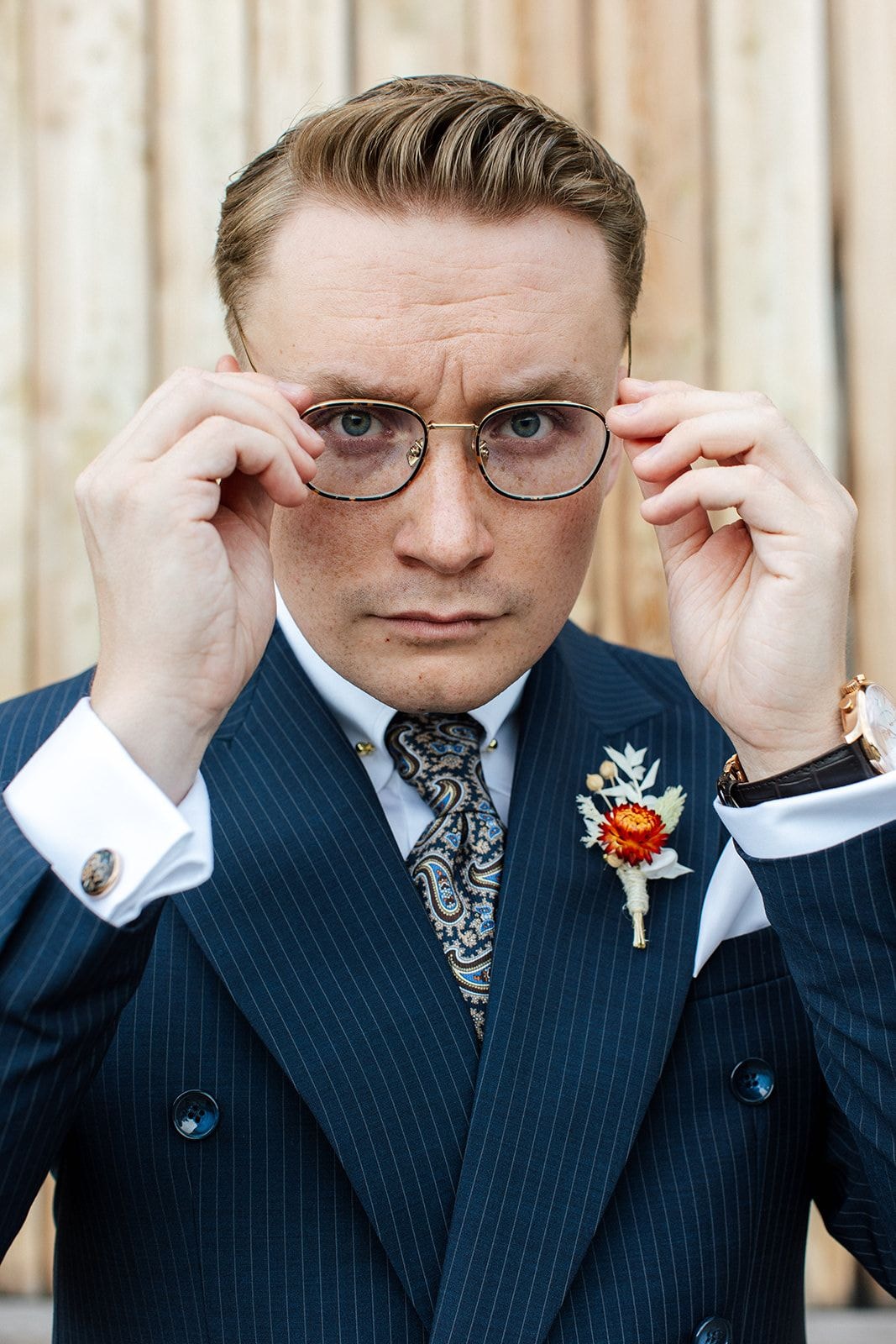 Mężczyzna w granatowym garniturze w paski, białej koszuli i paisley'owym krawacie poprawia okulary. Złota obrączka na palcu, broszka na klapie.