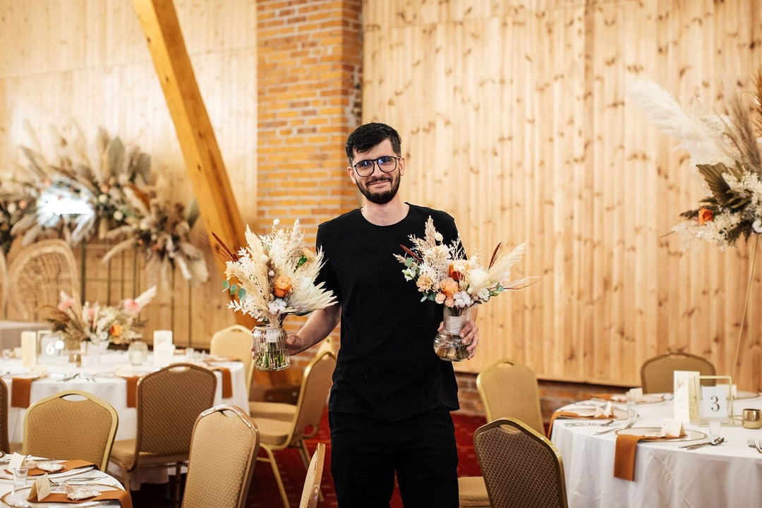 Florysta w czarnej koszulce trzyma dwie kompozycje kwiatowe w szklanych wazonach na tle drewnianej sali weselnej z udekorowanymi stołami.