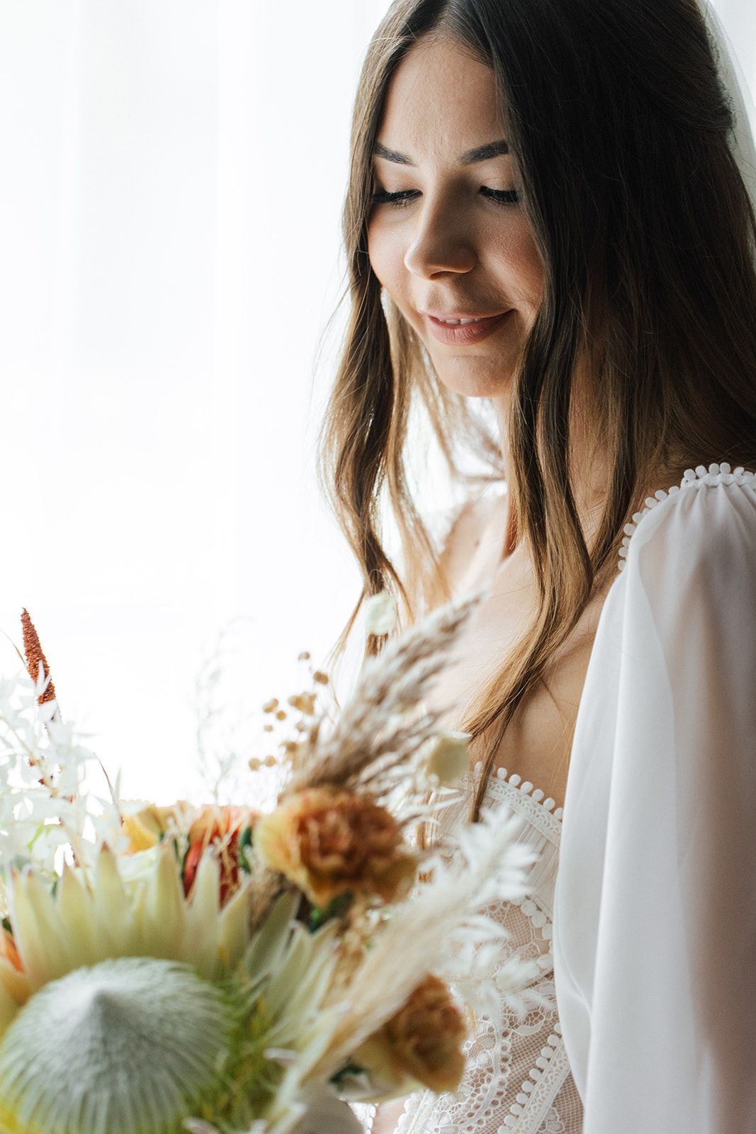 Kobieta w białej sukni ze złożonymi rękami patrzy na bukiet ślubny z suchymi kwiatami i trawami w jasnej palecie kolorów.