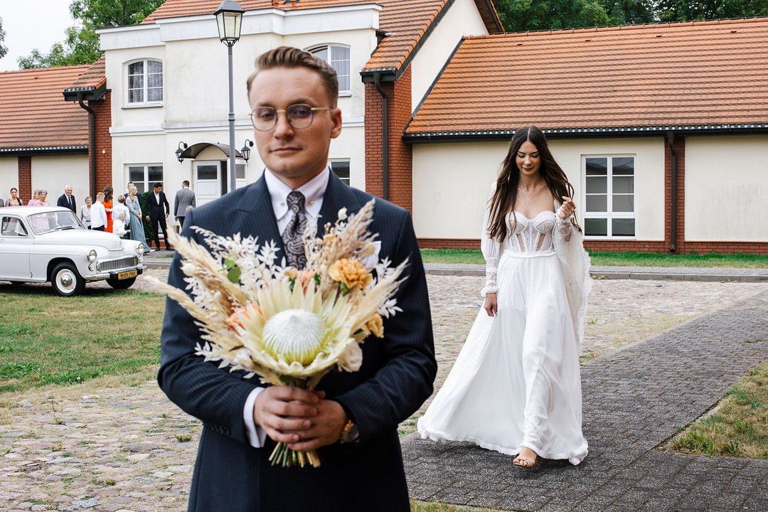 Mężczyzna w garniturze trzyma bukiet ślubny z jasnych kwiatów i zieleni, w tle kościół i goście