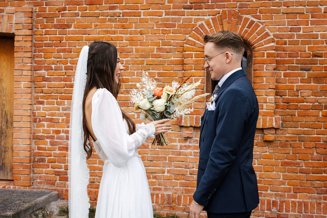 Para młoda przy ceglanej ścianie, kobieta w białej sukni ślubnej z welonu trzyma bukiet ślubny z białymi i pomarańczowymi kwiatami