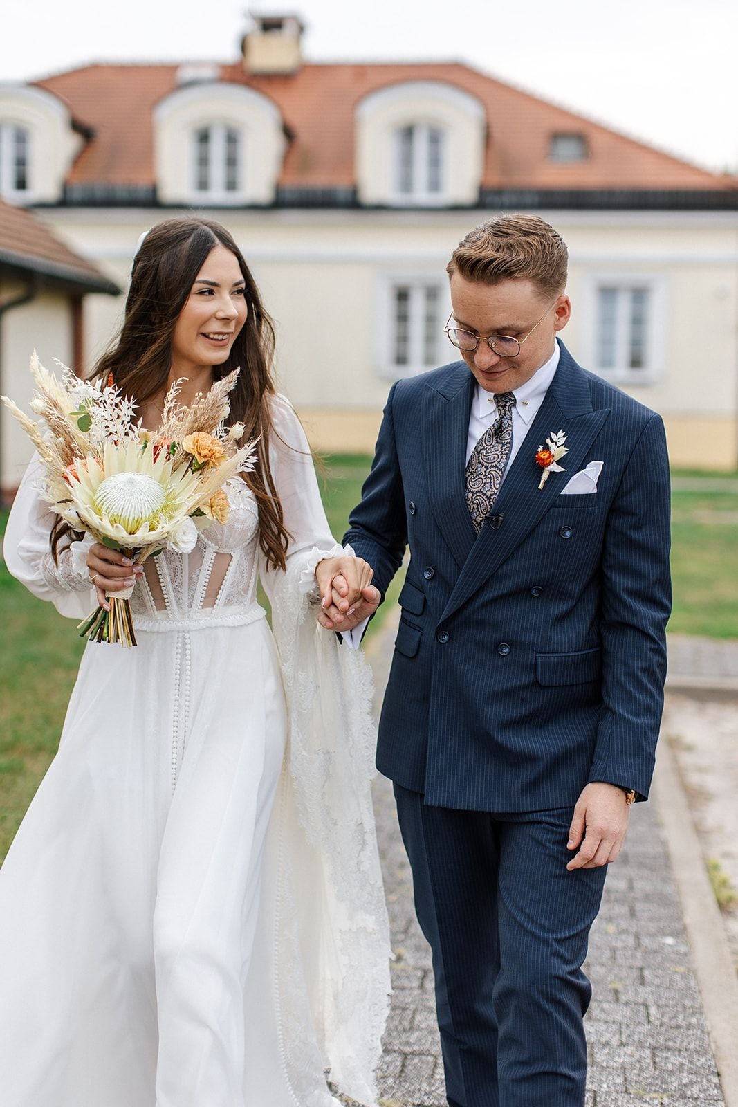 Para Młoda trzymająca się za ręce, kobieta w białej sukni ślubnej z bukietem kompozycja kwiatowa z suchymi kwiatami, mężczyzna w granatowym garniturze, tło zabytkowy pałac