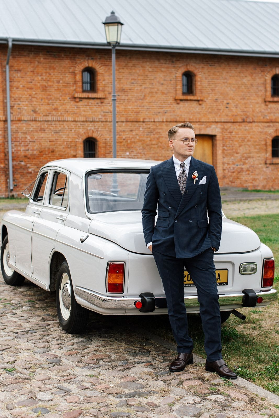 Mężczyzna w granatowym garniturze z butonierką stojący przy białym zabytkowym samochodzie przed ceglanym budynkiem