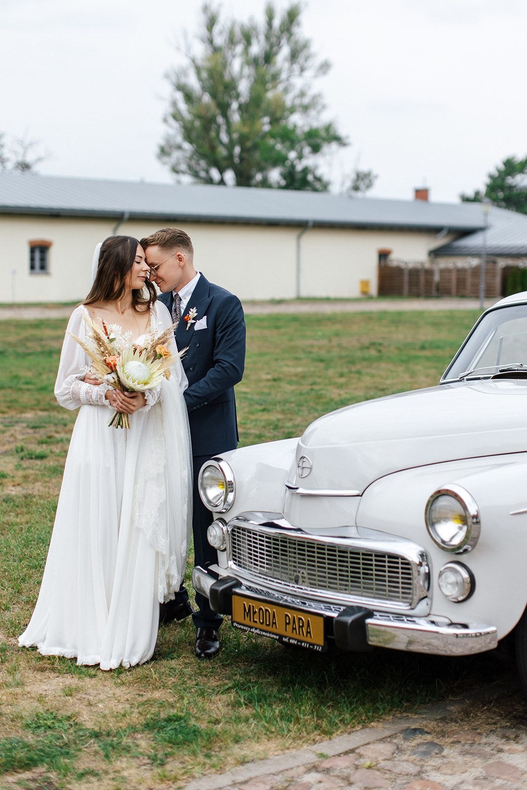 Para Młoda przy zabytkowym samochodzie, kobieta w białej sukni ślubnej trzyma bukiet ślubny w jasnej palecie, mężczyzna w granatowym garniturze, tło z zielenią i budynkiem
