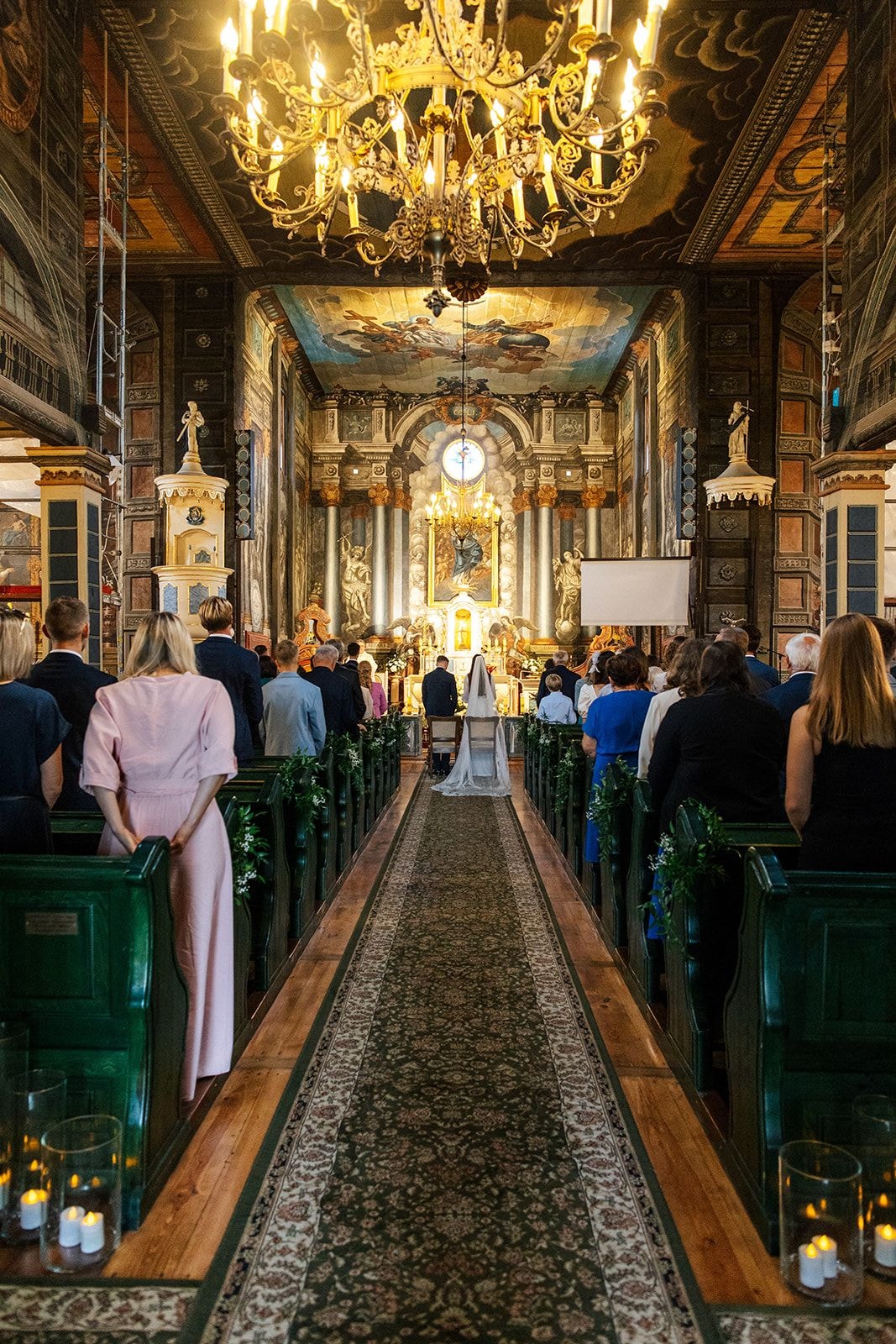 Wnętrze kościoła z dekoracją sali weselnej, złoty żyrandol, zielone girlandy wzdłuż alejki, goście siedzą w ławkach, ołtarz z kwiatami w tle.
