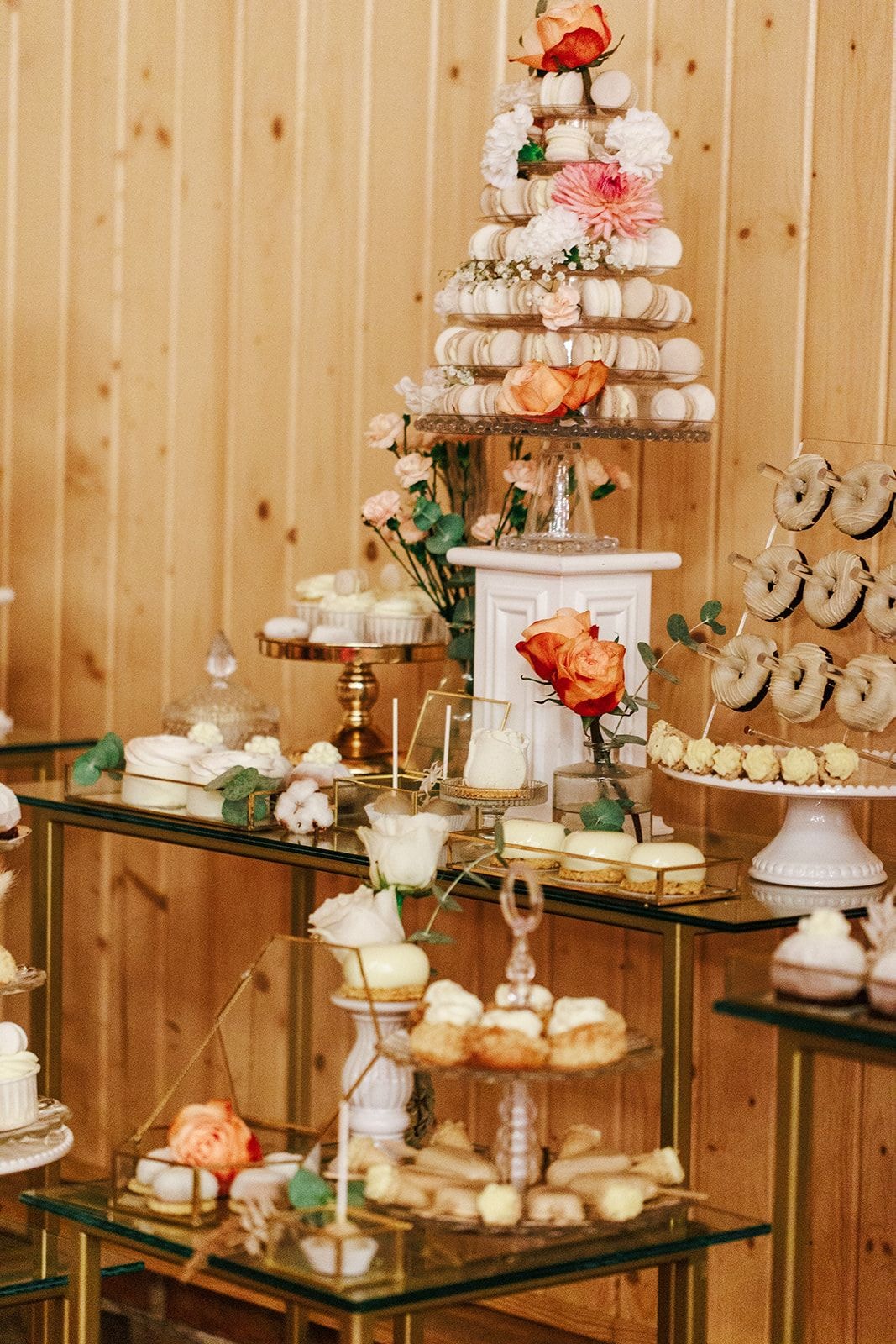 Dekoracja stołu z trzema poziomami makaroników, białymi kwiatami, różowymi różami i zielonymi akcentami na drewnianym tle sali.