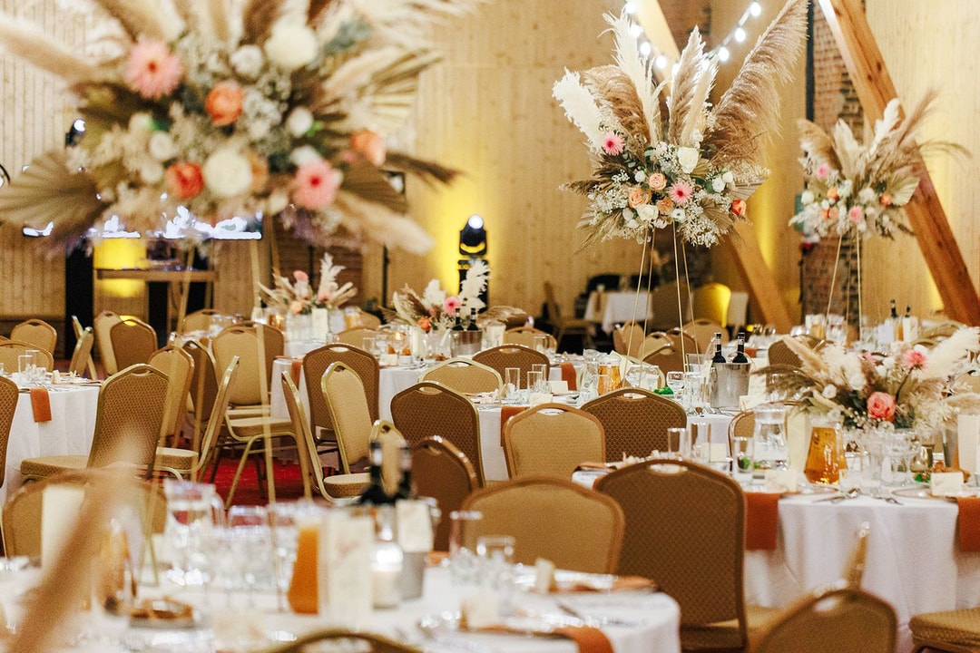 Aranżacja sali weselnej z białymi okrągłymi stołami, złotymi krzesłami i wysokimi kompozycjami kwiatowymi na drewnianych stojakach