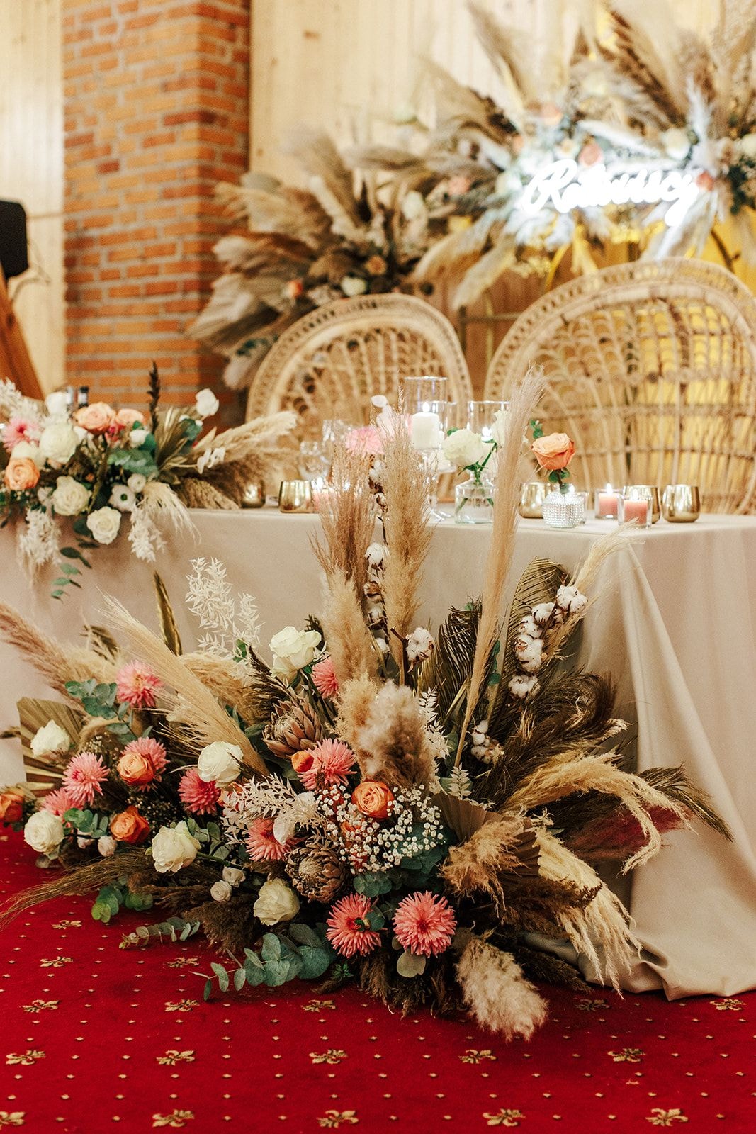 Dekoracja stołu z dużą kompozycją kwiatową na czerwonym dywanie, kwiaty w jasnej i różowej palecie, pampas grass, wianki słomiane w tle