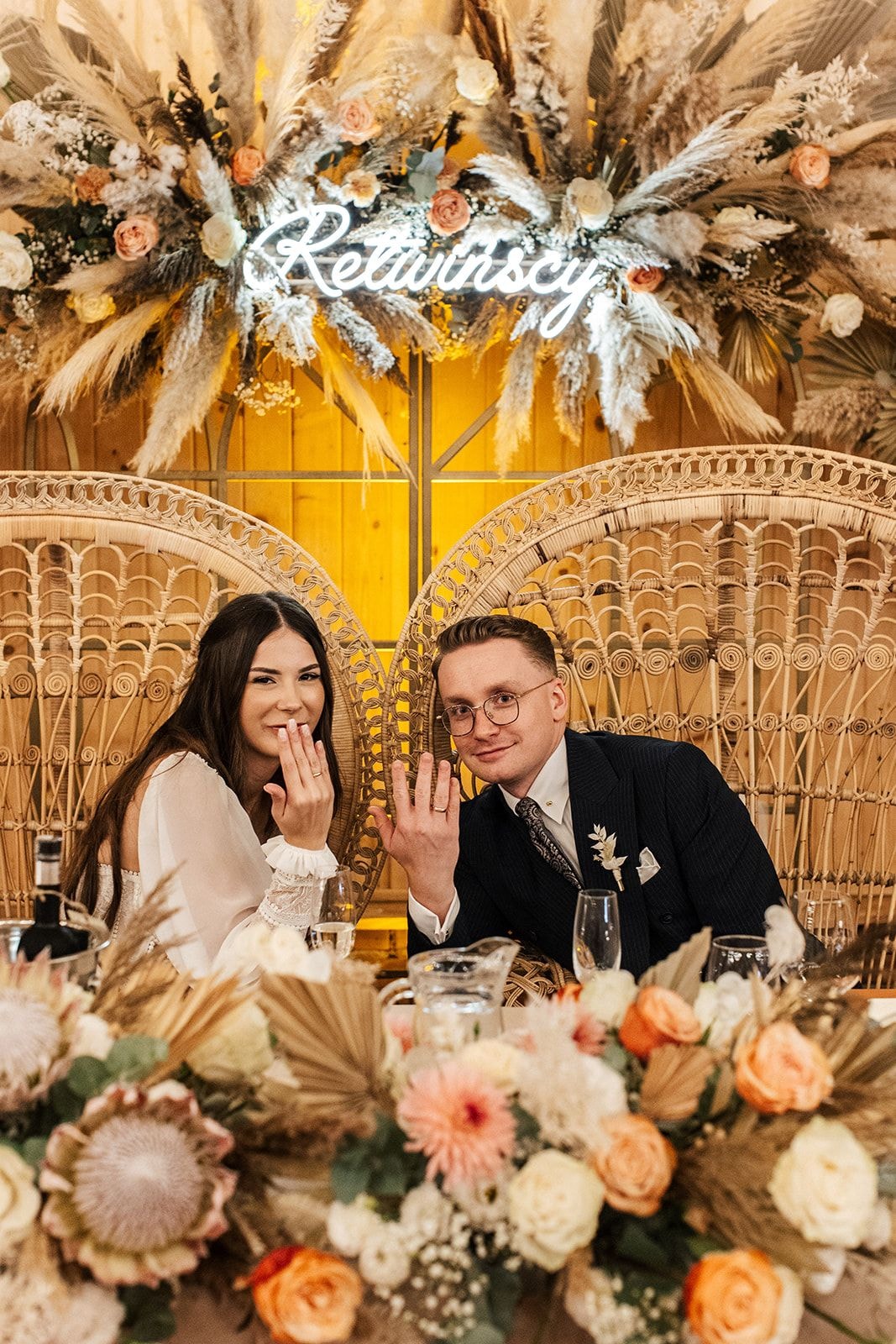 Para Młoda przy stole weselnym z dekoracją kwiatową. Złote liście palmowe, kwiaty w jasnej palecie, drewniany łuk i żółte tło za nimi.