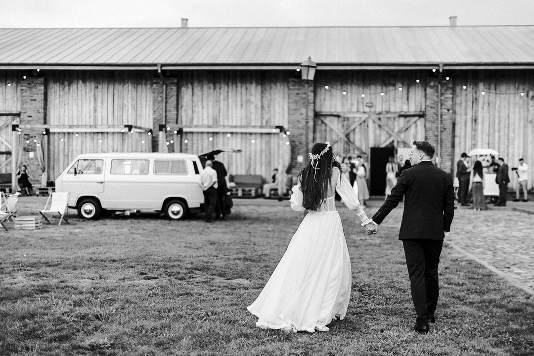Para młoda trzymająca się za ręce na trawie przed zabytkową stodołą, goście na tle, biały samochód, dekoracje sali weselnej widoczne w głębi.
