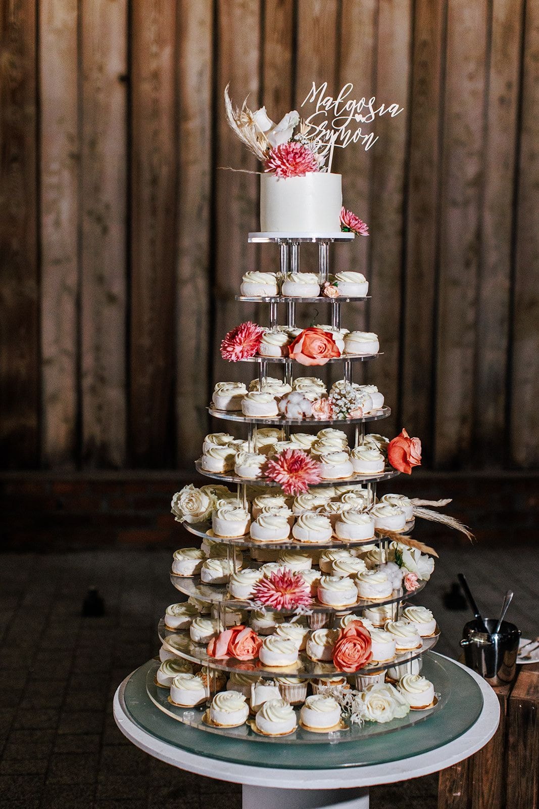 Piętrowa dekoracja stołu weselnego z białym tortem, makaronami oraz różowymi i pomarańczowymi kwiatami na tle drewnianych desek.