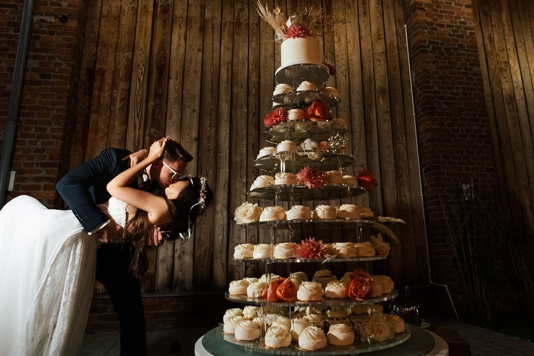 Para Młoda w drewnianej sali, mężczyzna w garniturze całuje pannę młodą w białej sukni przy piętrowej dekoracji sali weselnej z kwiatami.