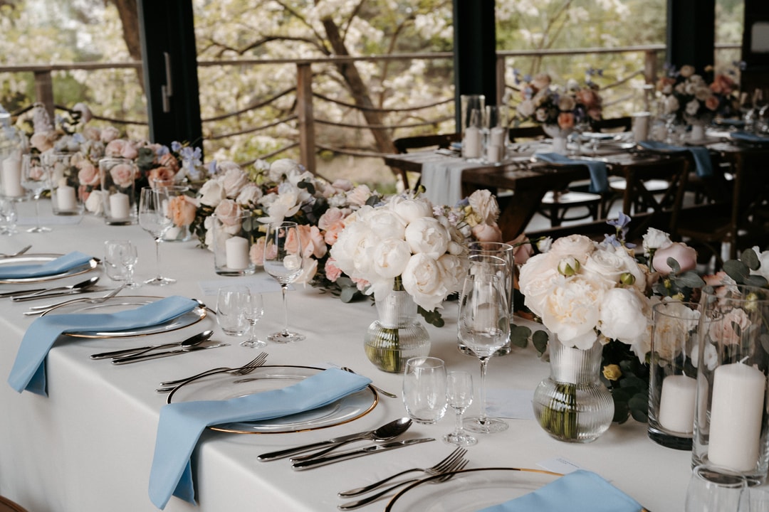 Aranżacja stołów weselnych z białymi peonią i różami w szklanych wazonach, niebieskie serwetki, sztućce i szklanki, tło z okien na ogród
