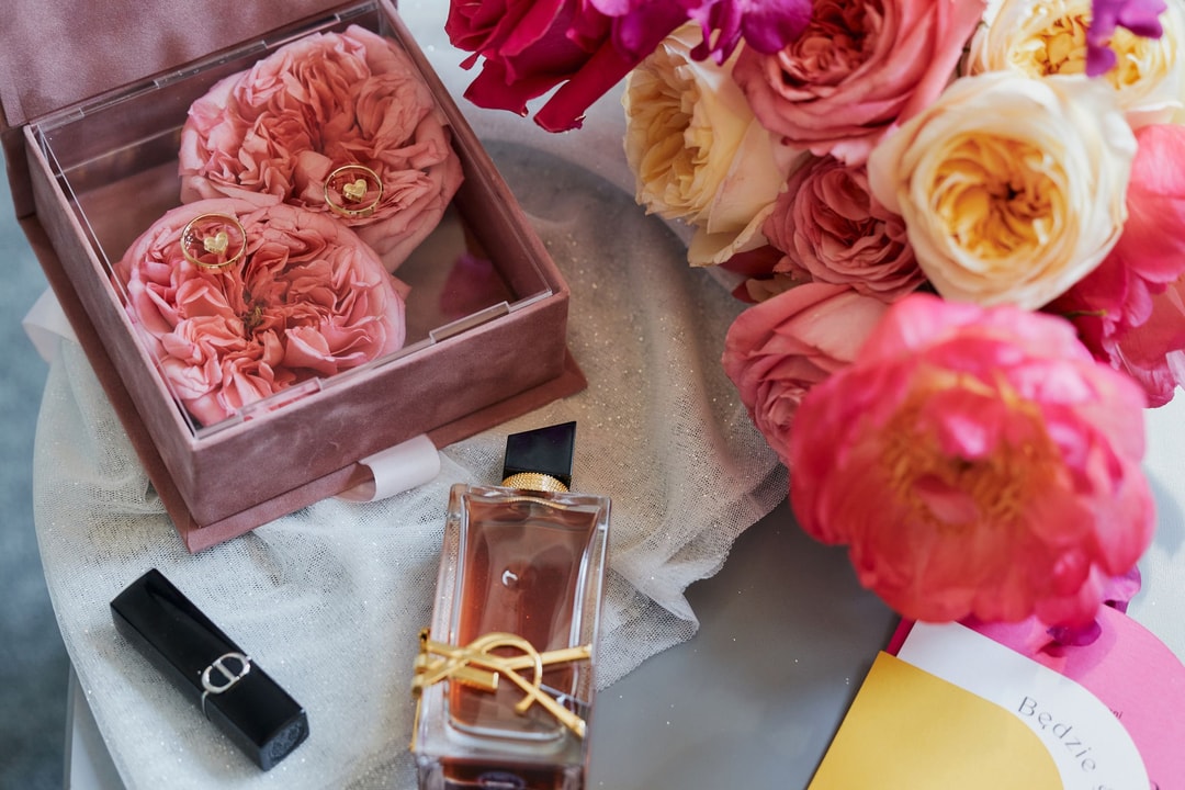 Kompozycja kwiatowa na stół w pudełku z różowymi kwiatami i złotymi pierścionkami, obok perfumy i kosmetyk