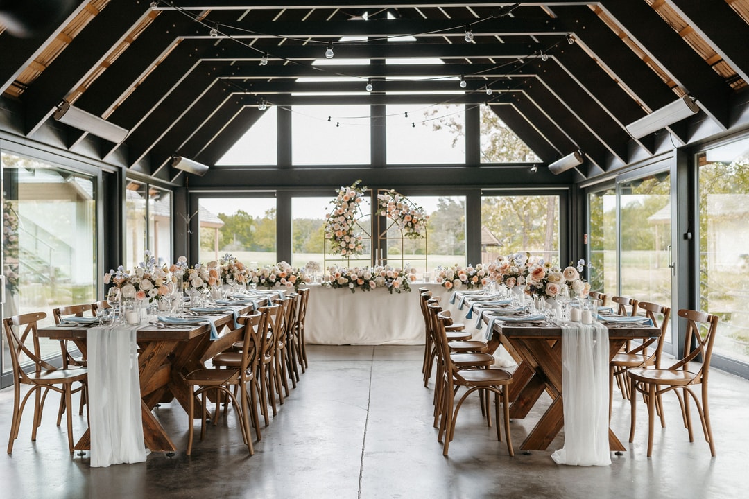 Dekoracje sali weselnej z dwoma długimi stołami, drewnianymi krzesłami, białymi obrusami i rozstawionymi kompozycjami kwiatowymi w jasnej palecie barw.