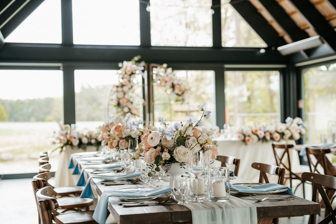 Aranżacja stołów weselnych z композycjami kwiatowymi w jasnej palecie, drewnianymi krzesłami i widokiem na jezioro przez panoramiczne okna sali.
