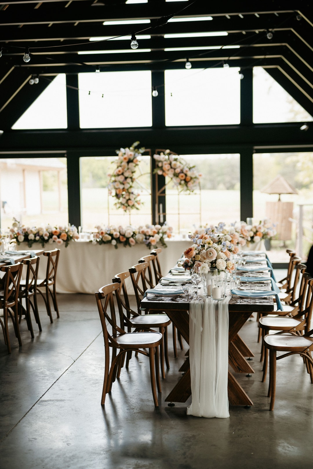 Dekoracje sali weselnej: długie stoły ze stołkami drewnianymi, białe bieżniki, kompozycje kwiatowe, okna z jasnym światłem
