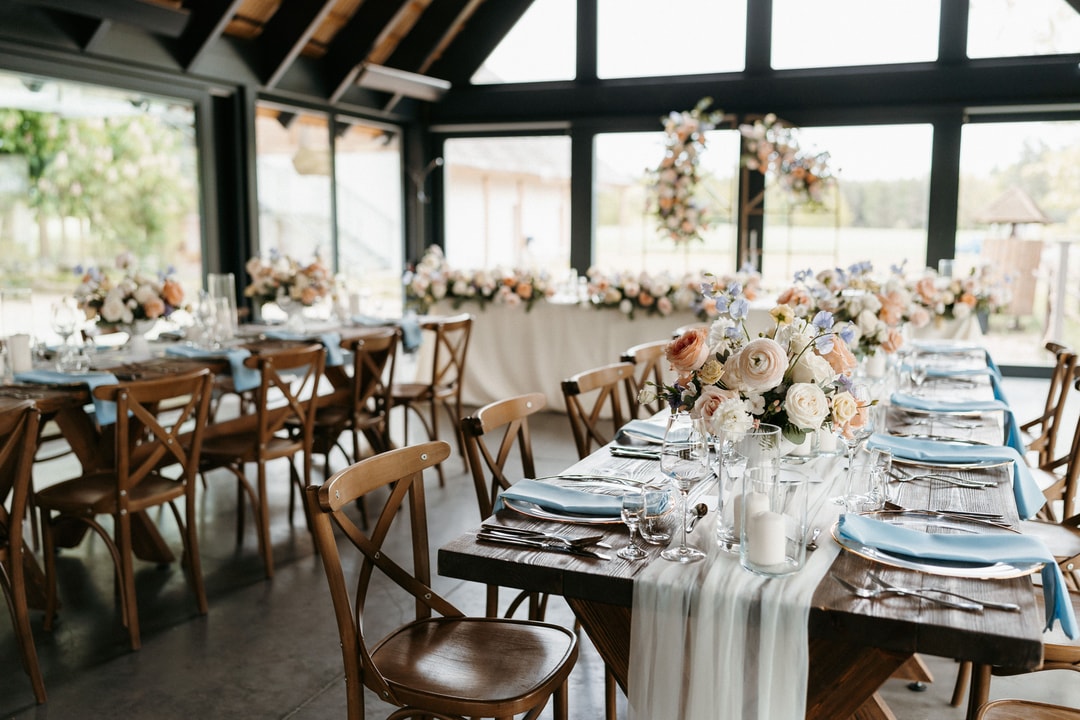 Elegancka aranżacja stołów weselnych w jasnej palecie z naturalnymi kompozycjami kwiatowymi, drewnianymi krzesłami i błękitnymi serwetkami w nowoczesnej sali.