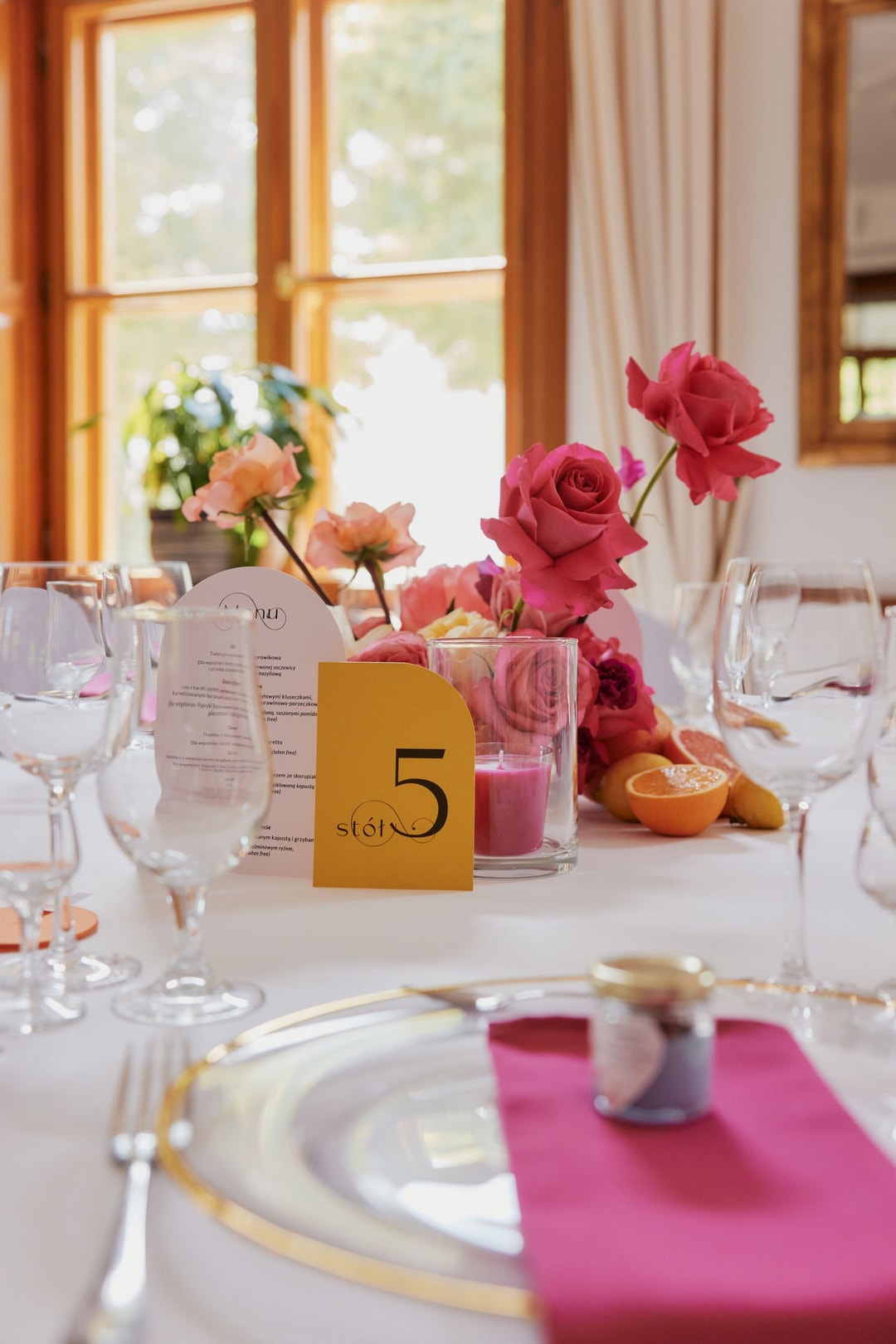 Dekoracja stołu z różowymi różami, kwiatami w jasnej palecie i złotym numerem stoła. Drewniane okna w tle.