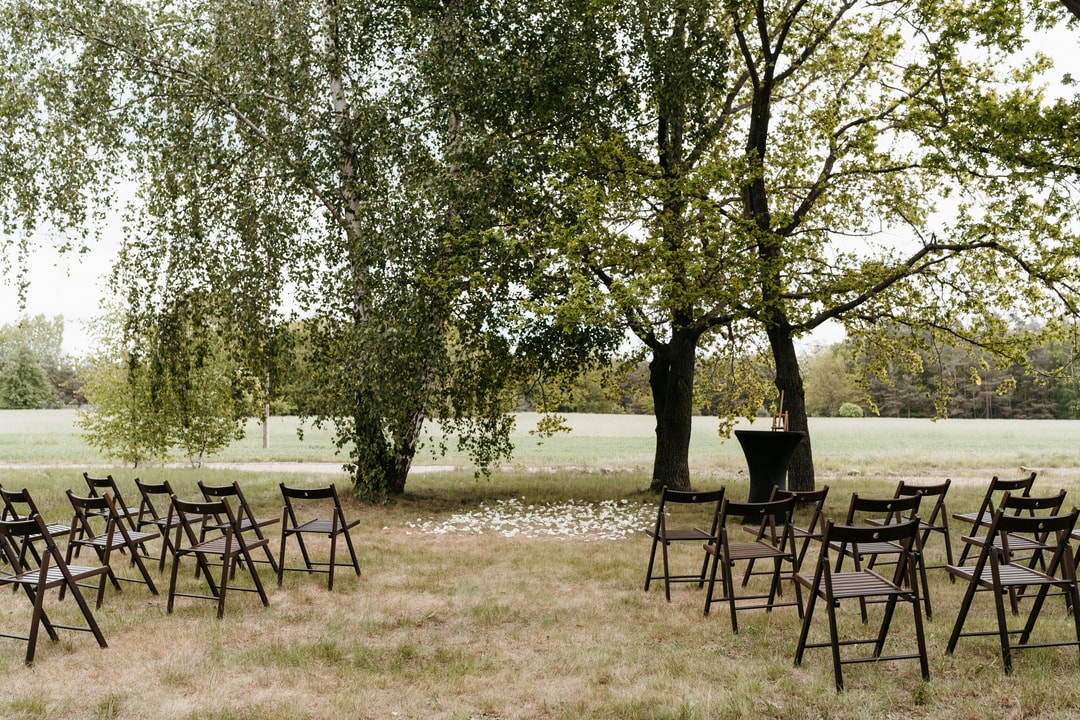Czarne krzesła ustawione na trawie pod drzewami, aranżacja stołów weselnych na łonie natury przy wodzie.