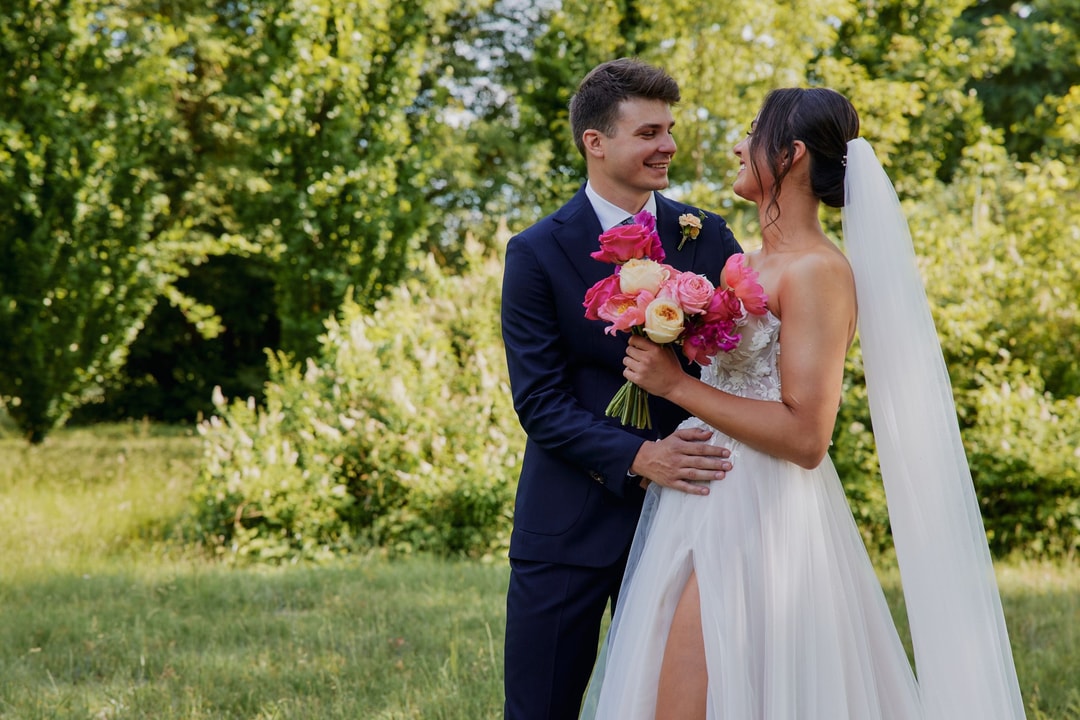 Para Młoda trzymająca bukiet ślubny z różowymi i śmietankowymi kwiatami na tle zieleni. Panna młoda w białej sukni, Pan Młody w ciemnoniebieskim garniturze.