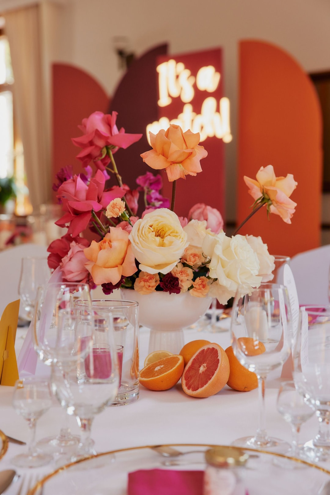 Kompozycja kwiatowa na stół w białej wazie z różami, kwiatami w odcieniach brzoskwini i głębokim różu, przy dekoracyjnym tle z napisami.