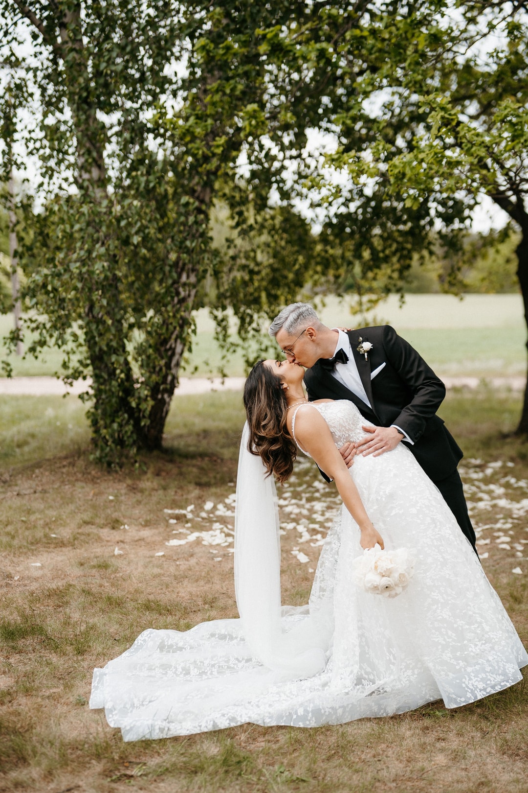 Para Młoda całuje się pod wierzbiną, panna młoda w białej sukni ślubnej z bukietem, pan młody w czarnym smokingu, tło park.