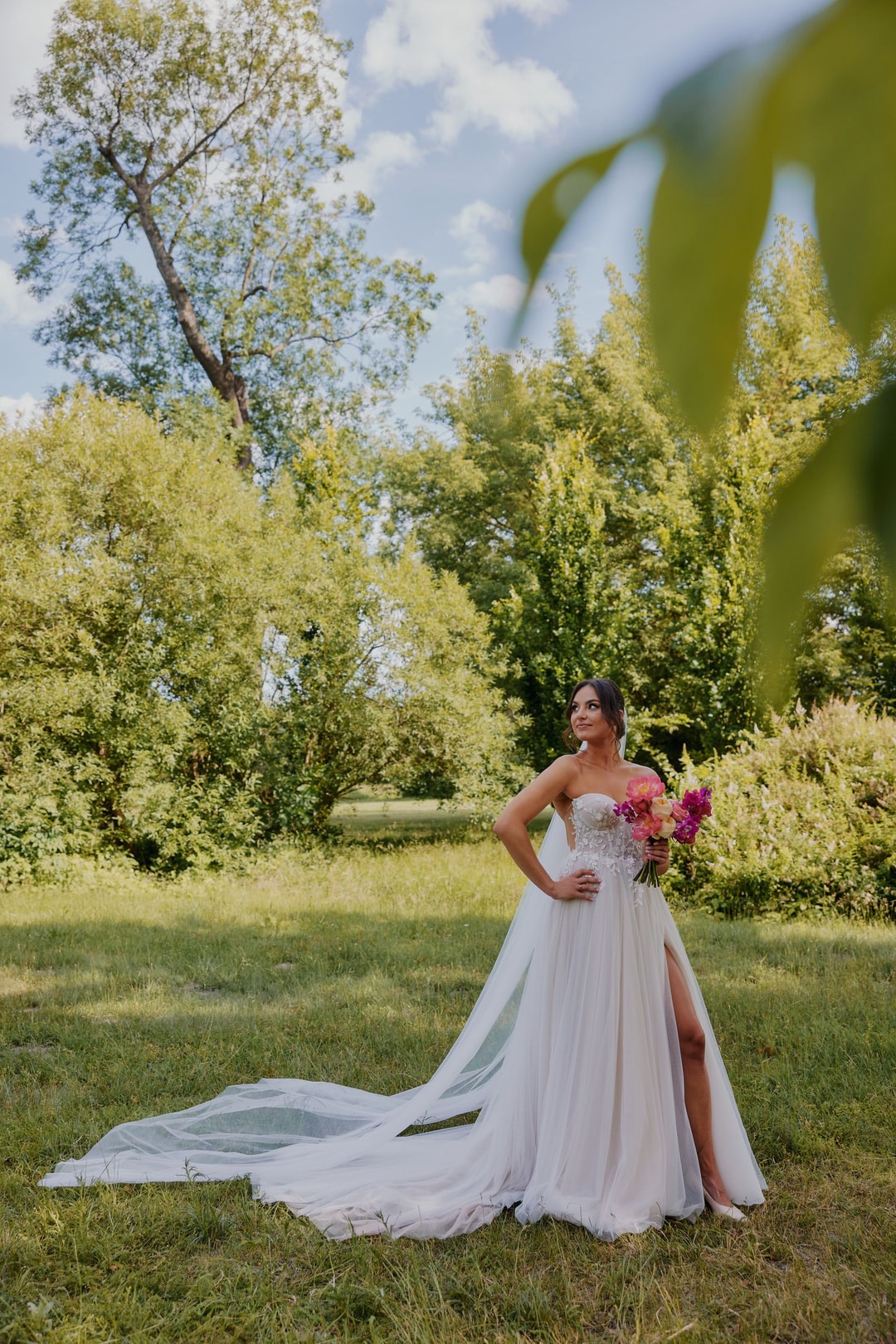 Kobieta w białej sukni z bukietem ślubnym stoi na łące z zielenią w tle, trzymając różowe i fioletowe kwiaty.