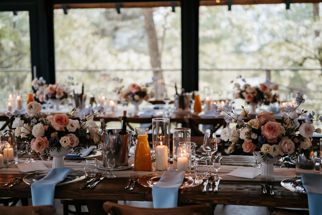 Aranżacja stołów weselnych z kwiatowymi kompozycjami w jasnej palecie, świecami i niebieskimi serwetkami przed panoramicznymi oknami