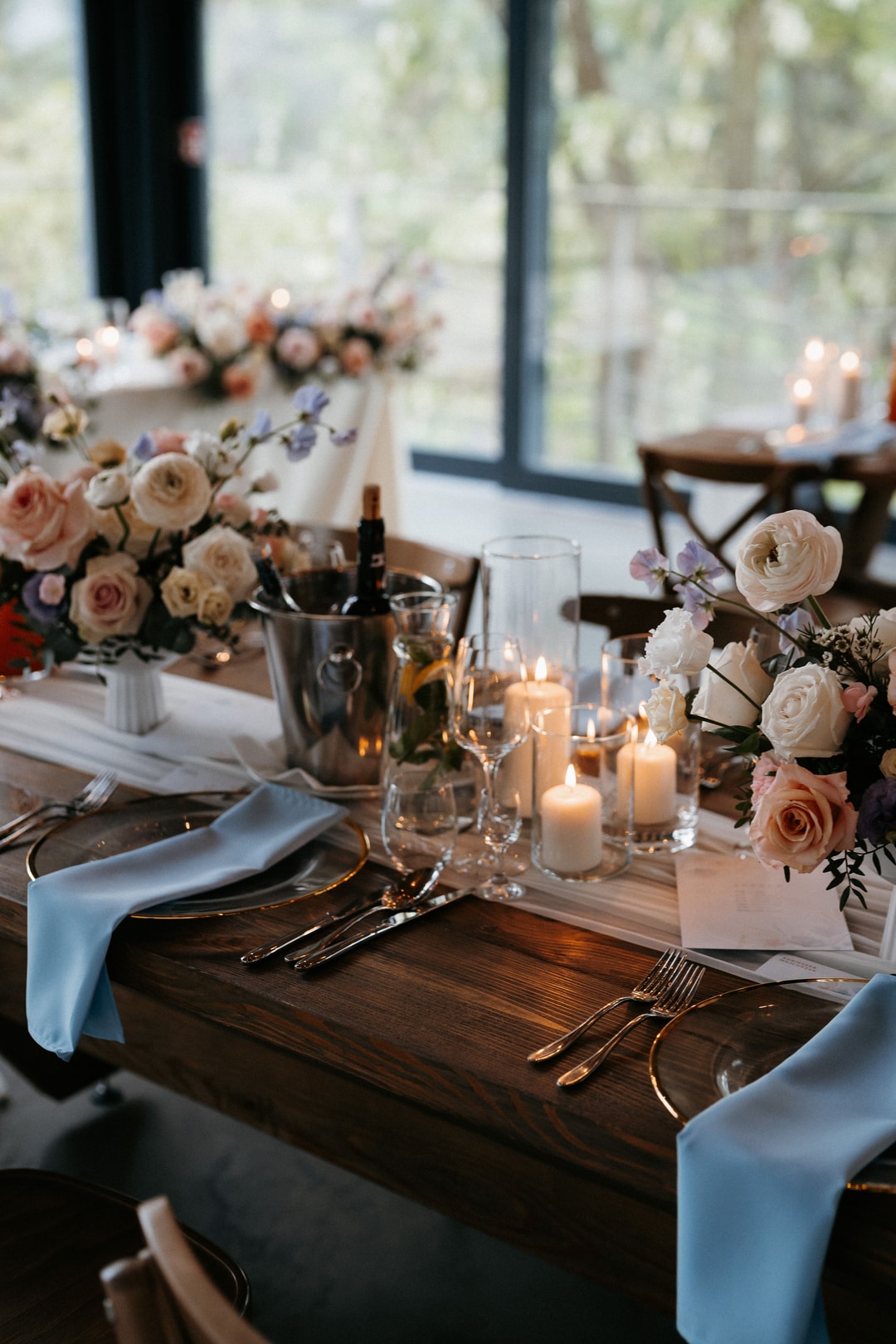 Dekoracja stołu z kompozycją kwiatową w jasnej palecie, świecami w szklanych lampionach, białą serwetką i sztućcami na drewnianym blacie