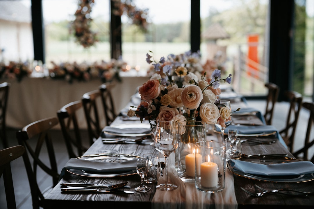 Aranżacja stołów weselnych z kompozycjami kwiatowymi w jasnej palecie, świeczkami i nakryciami w czarnym kolorze, w nowoczesnej sali z dużymi oknami