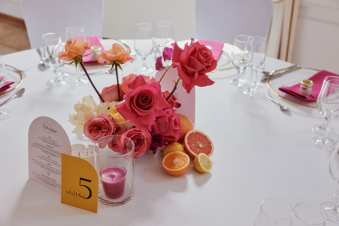 Kompozycja kwiatowa na stół z różami w odcieniach czerwieni i łososia, cytrusami oraz świecą zapachową, na białym obrusie