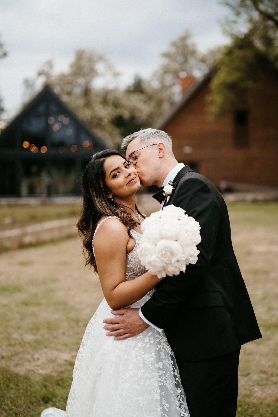 Para Młoda w strojach ślubnych trzymająca bukiet ślubny z białych kwiatów na tle zabudowań i zieleni.