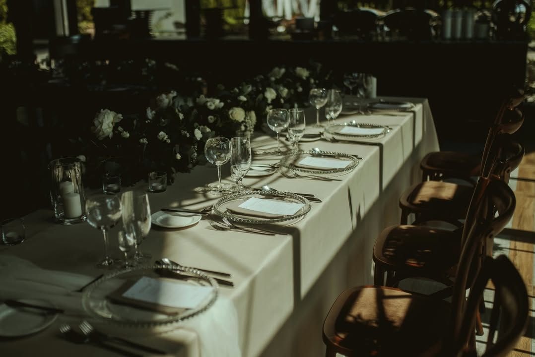 Aranżacja stołów z białym obrusem, białymi talerzykami, szklankami i sztućcami. Dekoracje sali weselnej z białymi kwiatami w wazach wzdłuż stołu. Drewniane krzesła po bokach.