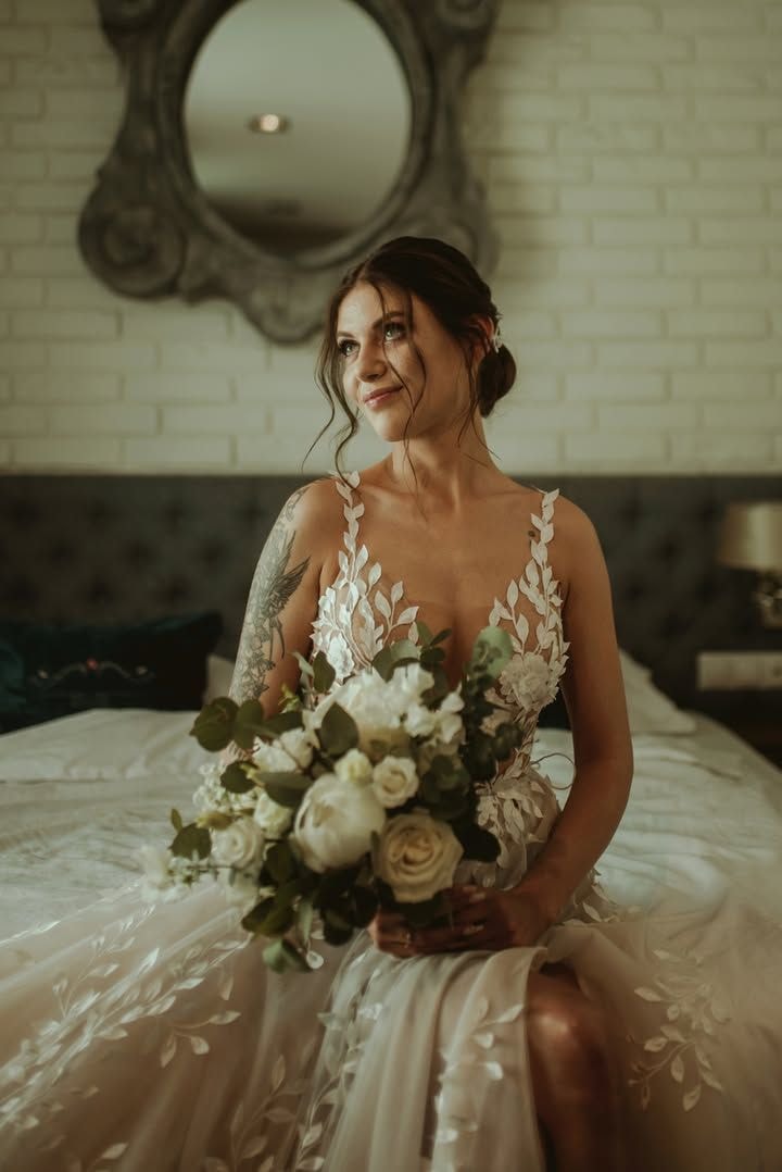 Kobieta w białej sukni z bukietem ślubnym zawierającym białe róże i zieloną oprawa, siedzi przy stole na tle ornamentacyjnego lustra