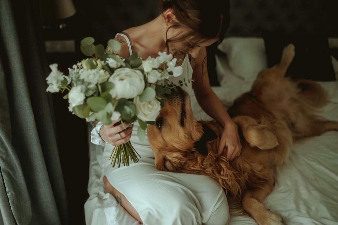 Kobieta w białej sukni trzymająca bukiet ślubny z białymi kwiatami i zielenią, obok brązowy pies