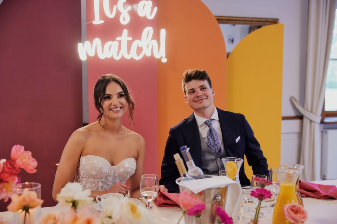 Panna młoda w białej sukni z bukietem ślubnym, róże w odcieniach różu i magenty, pan młody w granatowym garniturze, tło z białymi oknami.